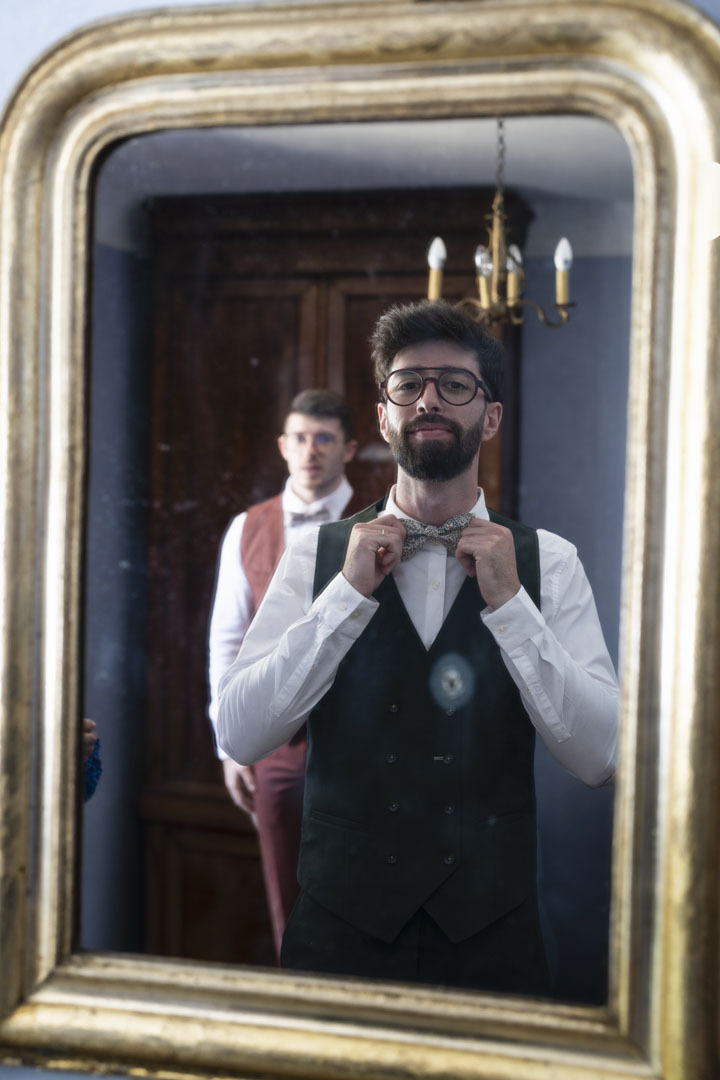 Photo des préparatifs du marié à la maison Lomprère , pau pyrénes atlantique. Le marié se regarde dans le miroir en se mettant correctement son noeud de papillon, pendant qu'un témoin le regarde en arrière plan. Cette photo est créative et à un coté ancien, car le miroir est ancien.